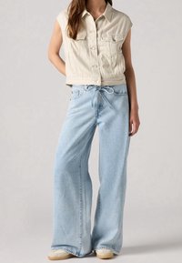 Beige kort denimväst med två bröstfickor, ihopparad med ljusblå vida jeans med dragsko i midjan och blek finish.