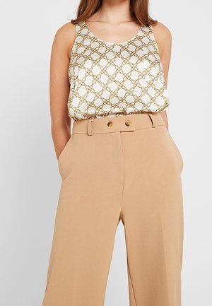 Kobieta ubrana w bezrękawnik o białym kolorze z wzorem złotego łańcucha, włożony w spodnie o wysokim stanie i beżowym kolorze z podwójnym zapięciem na guziki w talii.