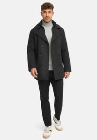 Schwarzer Wollmischungs-Peacoat mit doppelreihigen Knöpfen, steigendem Revers, kombiniert mit einem grauen Rollkragenpullover und schwarzen Hosen. Turnschuhe mit leichten Sohlen.