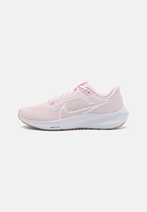 Zapatilla de running Nike de color rosa claro con el símbolo swoosh blanco, parte superior de malla, suela blanca acolchada y suela exterior de goma marrón sobre un fondo blanco.