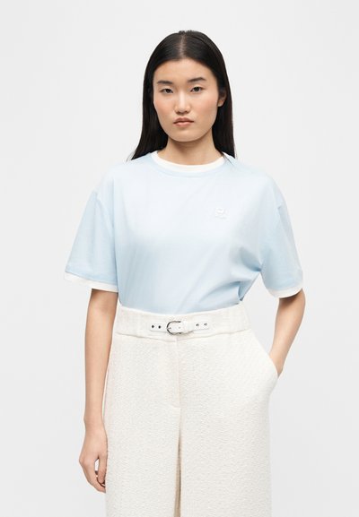 Νεαρή γυναίκα με μακριά σκούρα μαλλιά, φορώντας ένα ανοιχτό μπλε κοντομάνικο πουκάμισο, μπουσουλιασμένο μέσα σε ψηλόμεσα λευκά παντελόνια με υφή και ζώνη.