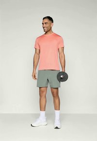Menns korallfarge atletisk T-skjorte og grønne shorts med tekstur, holder en svart vektplate, komplementert av hvite joggesko og sokker.