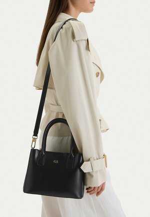 Donna con trench beige che porta una borsa a mano in pelle nera con logo dorato e tracolla rimovibile.