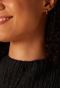 Boucles d'oreilles en or en forme de créole, ornées de pierres claires et noires, avec une surface texturée et un petit design moderne, portées par une personne aux cheveux bouclés et foncés.