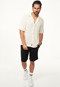 Kurzärmliges, cremefarbenes Strickshirt mit strukturiertem Muster, kombiniert mit schwarzen Shorts und weißen Sneakers mit schwarzen Streifen auf den Socken.