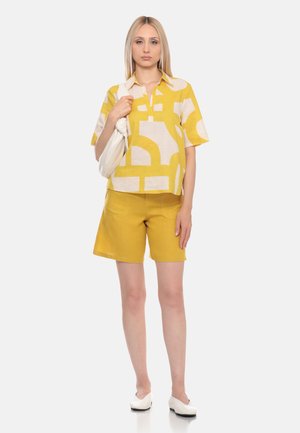 Donna in piedi che indossa una camicia gialla a maniche corte fantasia e pantaloncini gialli tinta unita, tenendo una borsa a tracolla bianca, con scarpe bianche su sfondo chiaro.