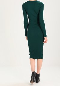 Femme portant une robe midi ajustée à manches longues en maille côtelée de couleur vert foncé et des bottines à talons noires, vue de dos.