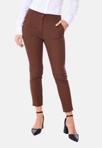 Pantaloni marroni attillati con una texture liscia, chiusura a bottone, tasche frontali e lunghezza cropped, abbinati a scarpe nere con tacco alto.