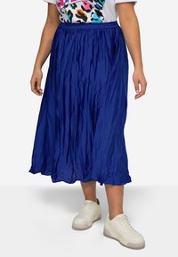Falda plisada azul de largo medio, hecha de una tela ligera y brillante. Presenta una cinturilla elástica y una silueta fluida. Se lleva con zapatillas blancas.