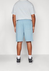 Muž v světle modrých šortkách, bílém a modrém proužkovaném tričku, bílých ponožkách a černých teniskách, stojící čelem ke jednoduché zdi.