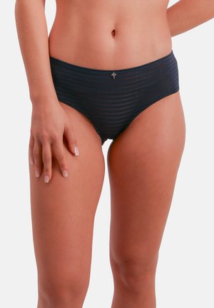 Femme portant un sous-vêtement bikini rayé bleu foncé, semi-transparent, montrant le torse de la taille jusqu'au milieu de la cuisse sur fond uni.