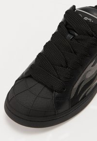 Sneaker nera con punta in gomma testurizzata, dotata di spesse stringhe nere e un design laterale in grigio scuro. Materiale superiore in pelle liscia.