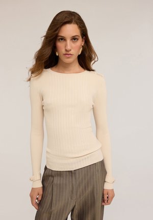 Junge Frau mit langen braunen Haaren, die einen beigen ribbeligen Strickpullover und braune Nadelstreifenhosen trägt, steht vor einem schlichten hellen Hintergrund.