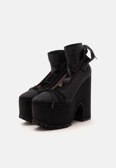 Paire de chaussures à talons hauts plateformes noires avec bouts arrondis, brides de cheville nouées en nœuds et talons blocs épais sur fond uni.
