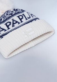 Berretto bianco lavorato a maglia con una fascia a motivo blu navy, caratterizzato da un pompon soffice in cima e una toppa rettangolare con una bandiera.