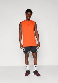 Oransje ermeløs skjorte med svart logo, kombinert med svarte mesh-shorts med hvite detaljer. Svarte joggesko med røde detaljer.