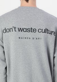 Sudadera gris con un gran gráfico de texto negro en la parte posterior que dice "no desperdicies cultura" y "MAISON D'ART".