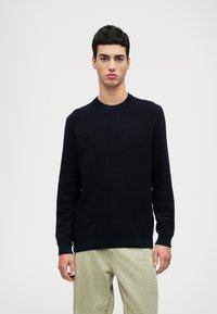 Μπλε πλεκτό πουλόβερ με στρογγυλή λαιμόκοψη, μακριά μανίκια, υφή υφάσματος και ραμμένες μανσέτες, συνδυασμένο με ανοιχτόχρωμες φαρδιές παντελόνες.