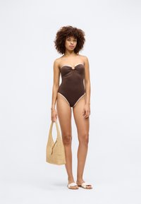 Brūns bezsniega viengabala peldkostīms ar baltu apdari, mezglu detaļu priekšpusē un augstiem izgriezumiem, kopā ar austas salmu somu un sandālēm.