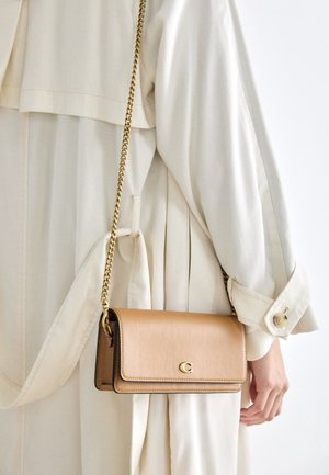 Person iført en cremefarvet trenchcoat, der holder en beige læder crossbody taske med en guldkæde og lille gyldent "C" logo.
