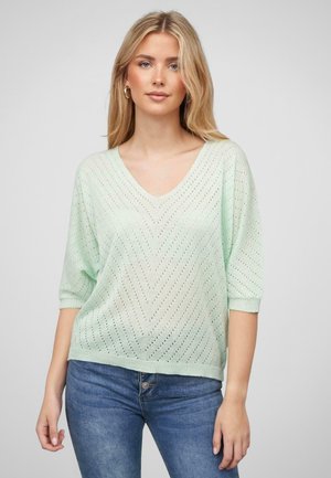 Mintfarbener Strickpullover mit V-Ausschnitt, kurzen Ärmeln und einem Chevron-Muster aus durchbrochener Gestaltung. Kombiniert mit blauen Jeans.