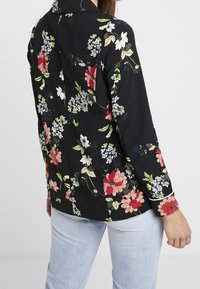 Blommönstrad jacka i svart, med färgglada blommor och blad. Långa ärmar, krage, slätt tyg; detaljer vid ärmsluten.