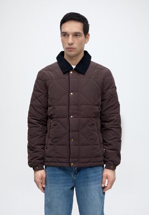 Homme portant une veste matelassée marron avec un col noir et un jean bleu, debout devant un fond clair uni.