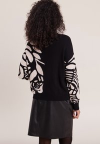 Schwarzer Pullover mit beigem Blumenmuster an den Ärmeln, mit lockerer Passform und gerippten Bündchen; kombiniert mit einem schwarzen Kunstlederrock.