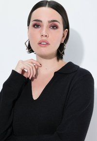 Czarny sweter z dzianiny z dekoltem w serek i prążkowanymi mankietami, zestawiony z dużymi, geometrycznymi srebrnymi kolczykami. Modelka ma krótkie ciemne włosy.