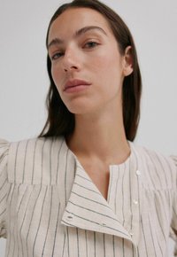 Lichtbeige overhemdd met blauwe strepen, korte pofmouwen en een puntige kraag. Bevat een zak en heeft een gladde textuur.