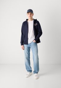 Un sweatshirt zippé bleu marine sur un t-shirt blanc, un jean bleu clair et des baskets blanches. Comprend une casquette bleue. Tenue décontractée avec un design minimal.