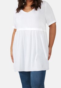 Vit kortärmad topp med V-ringning, samlad textur vid midjan och fickor, tillverkad av lätt material. Kombinerad med blå jeans.