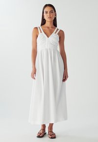 Hvid maxikjole med rynket overdel og tynde skulderstropper. Glat, let stof falder til gulvet. Bæres med sorte sandaler.