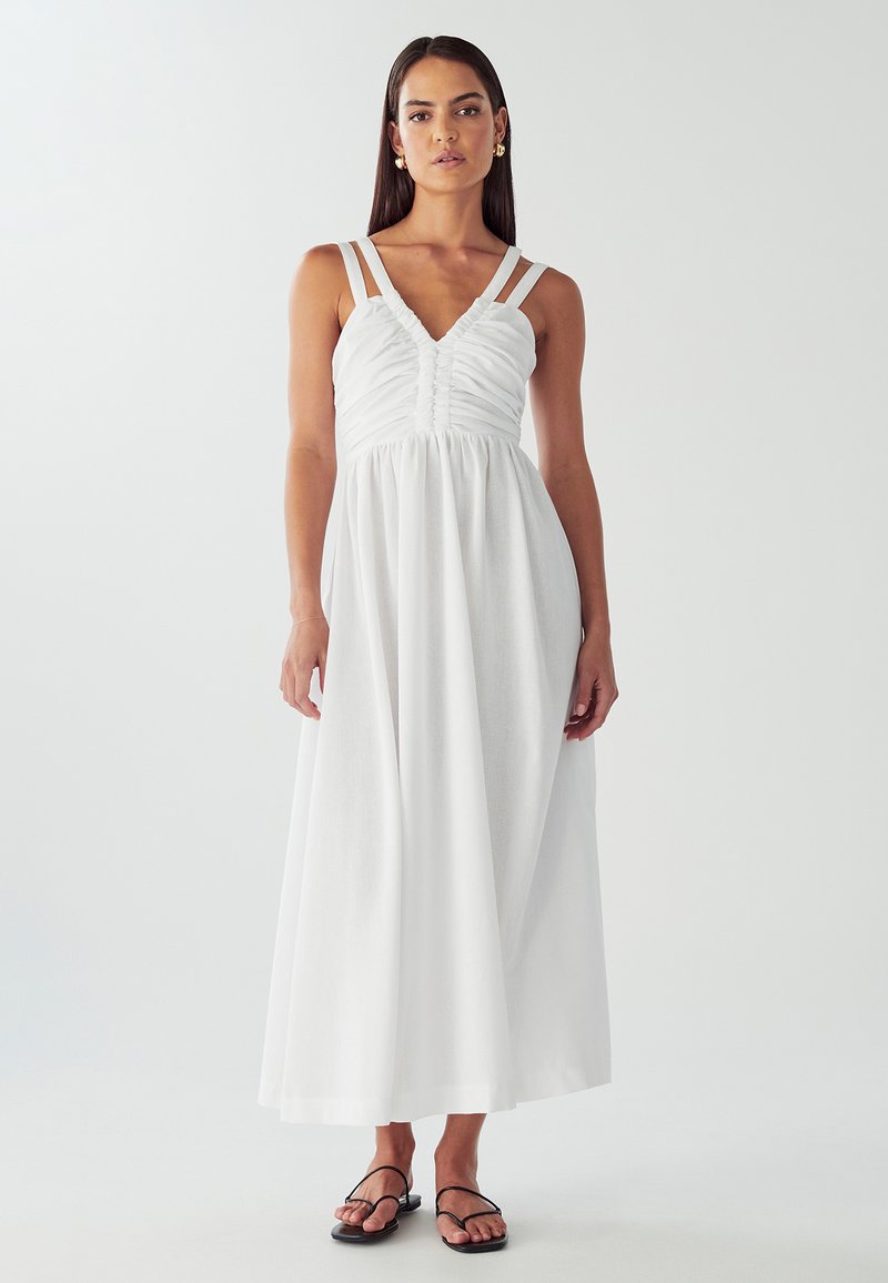 Hvid maxikjole med rynket overdel og tynde skulderstropper. Glat, let stof falder til gulvet. Bæres med sorte sandaler.