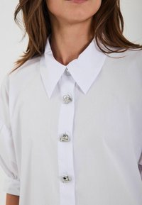 Camicia bianca con un colletto ampio, dettagli con bottoni argentati e una texture liscia. Le maniche a sbuffo aggiungono volume al design.