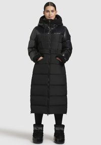Lang zwart pufferjack met een riem in de taille, een ritssluiting aan de voorkant en een oversized kraag. Kenmerkt zich door contrasterende glanzende en matte materialen, twee zijzakken.