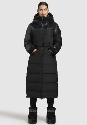 Lang zwart pufferjack met een riem in de taille, een ritssluiting aan de voorkant en een oversized kraag. Kenmerkt zich door contrasterende glanzende en matte materialen, twee zijzakken.