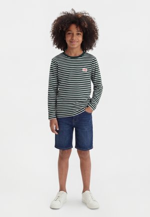 Enfant aux cheveux bouclés portant un t-shirt à manches longues rayé vert et blanc, un short en jean et des baskets blanches, debout avec une main dans la poche.