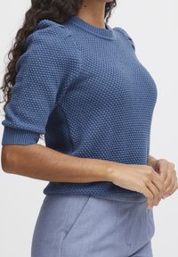 Modrý pletený svetr s texturovaným vzorem, krátkými rukávy a žebrovanými manžetami. Nošený s světle šedými kalhotami na míru.