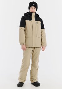 Beige und schwarze isolierte Jacke mit großen Taschen, einer Kapuze und passenden beigen Hosen. Das Outfit beinhaltet eine schwarze Mütze und robuste schwarze Stiefel.