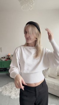 Bílá zkrácená mikina s žebrovaným lemem, doplněná černými kalhotami. Dlouhé blond vlasy částečně zasunuté za černou čelenku.