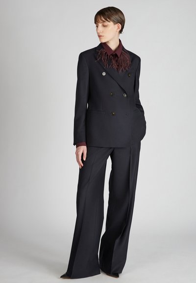 Abito nero doppiopetto con bottoni argentati, pantaloni a gamba larga e camicia bordeaux con colletto caratterizzata da un accento in piuma testurizzata.