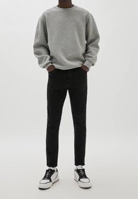 Personne portant un sweat gris, un pantalon slim noir et des baskets blanches avec des détails noirs, debout les mains dans les poches.
