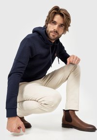 Marineblauwe getextureerde hoodie met een trekkoordcapuchon, beige slim-fit broek en bruine leren Chelsea boots met elastische zijpanelen.