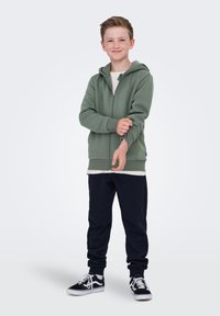 Niño con sudadera verde con cremallera subiendo la manga, vistiendo pantalones de chándal negros y zapatillas negras, de pie contra un fondo claro.