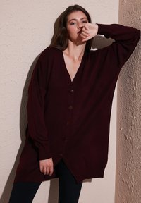 Sieviete burgundijas krāsas pārmērīgā pogājama džemperī balstās pret teksturētu bēšu sienu ar paceltu vienu roku, ģērbta melnās biksēs.