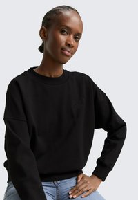 Donna con capelli intrecciati che indossa una felpa nera con un sottile ricamo "5th Ave Club", abbinata a jeans azzurri, posando con la mano sul fianco.