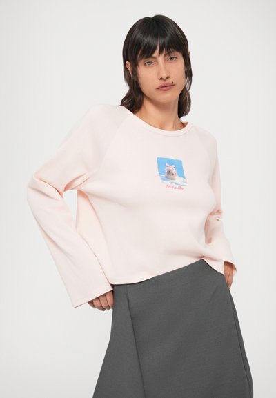 Világos rózsaszín hosszú ujjú pulóver grafikus nyomattal, amelyen egy masnit viselő nyúl látható. Laza fazonú és kerek nyakkivágású, szürke szoknyával párosítva.