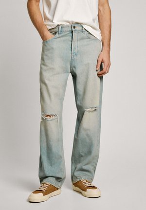 Jeans de mezclilla azul claro con un ajuste relajado, que presentan rodillas desgastadas, una cremallera y bolsillos laterales, combinados con zapatillas marrones.