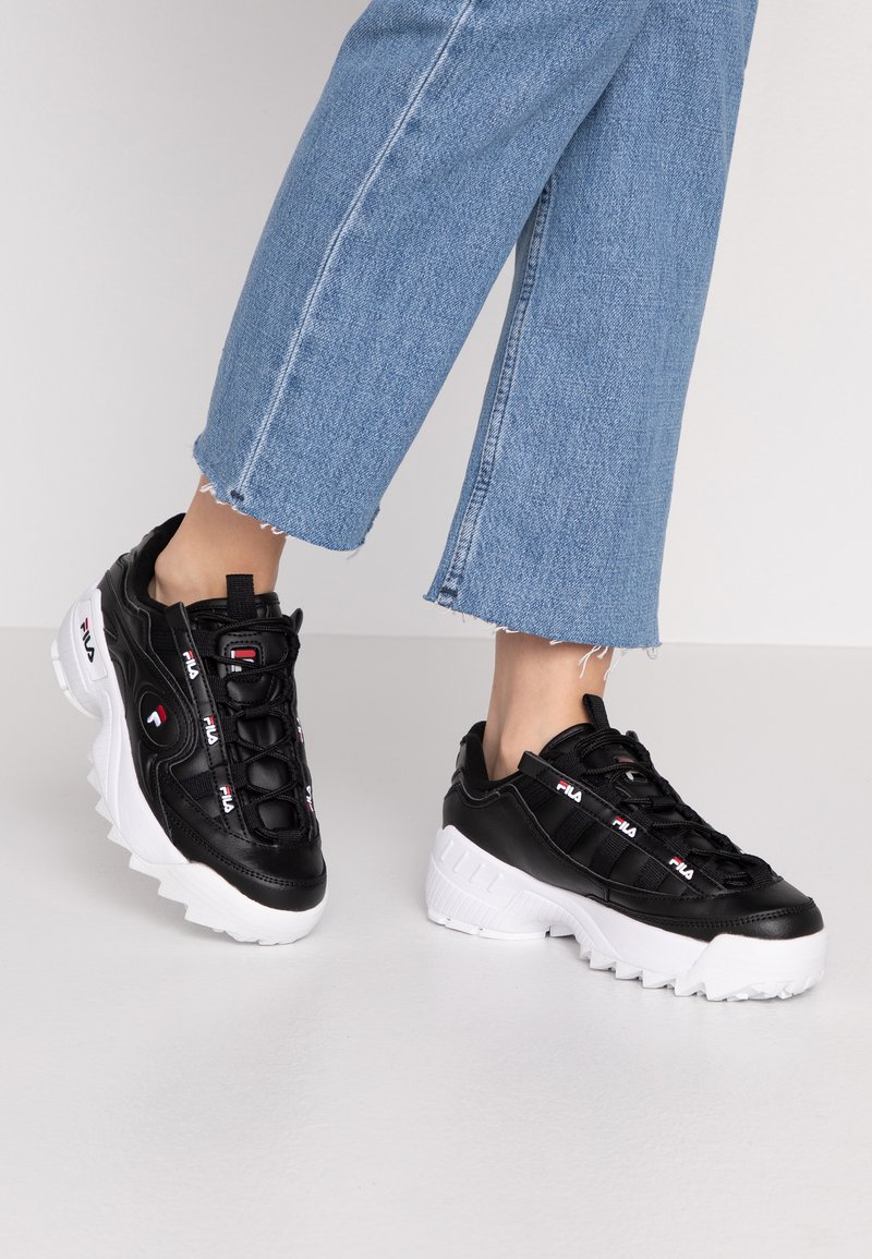 Czarne skórzane sneakersy z masywną białą podeszwą, z sznurówkowym zapięciem i logotypem Fila z boku. Dżinsowe spodnie z postrzępionymi mankietami.
